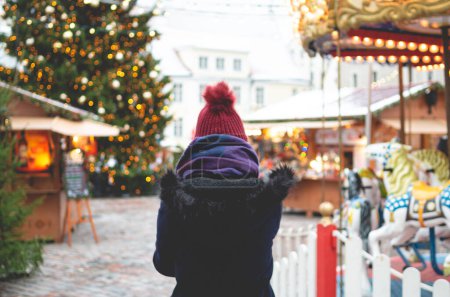 Téléchargez les photos : Une jeune femme vêtue d'un manteau d'hiver bleu et d'un chapeau rouge debout sur une place de l'hôtel de ville de Tallinn par une froide journée d'hiver avec un sapin de Noël et un repas de Noël en arrière-plan. - en image libre de droit
