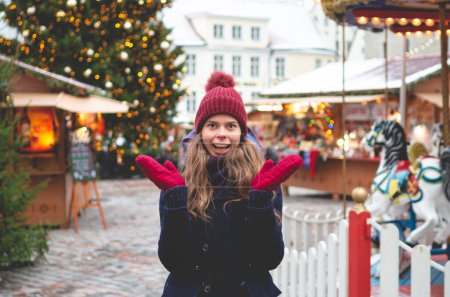 Photo for A handsome young woman in a blue winter coat, red hat and gloves making a surprise hand movement while standing on a Tallinn Town Hall Square on a cold winter day with a Christmas tree and a Christmas fare in the background. - Royalty Free Image