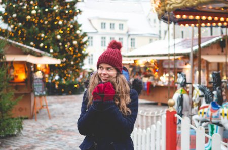 Photo for A handsome young woman in a blue winter coat, red hat and gloves holding hands together and standing on a Tallinn Town Hall Square on a cold winter day with a Christmas tree and a Christmas fare in the background. - Royalty Free Image