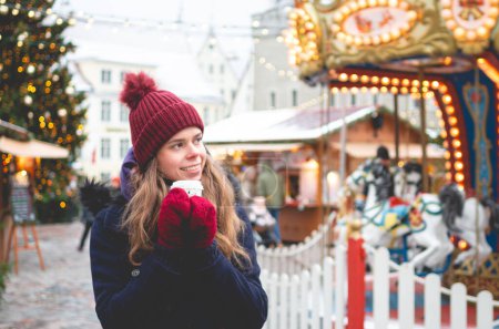 Photo for A handsome young woman in a blue winter coat, red hat and gloves holding coca cup and standing on a Tallinn Town Hall Square on a cold winter day with a Christmas fare and a children's carousel in the background. - Royalty Free Image