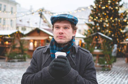 Téléchargez les photos : Un beau jeune homme en manteau gris avec une casquette écossaise en tweed et une écharpe en tartan tenant une tasse de cacao et debout sur une place de l'hôtel de ville de Tallinn par une journée d'hiver lumineuse avec un arbre de Noël et un repas de Noël en arrière-plan. - en image libre de droit