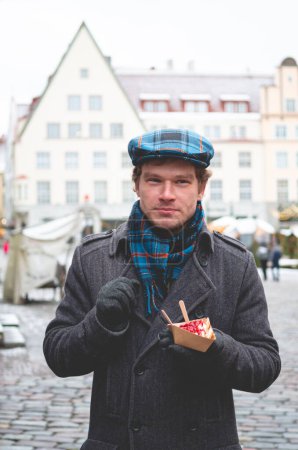 Photo for A handsome young man in a grey coat with a Scottish tweed cap and tartan scarf holding a dessert and standing on a Tallinn Town Hall Square on a bright winter day with a Christmas fare in the background. - Royalty Free Image