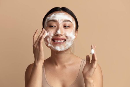 Foto de Cuidado de la piel facial. Mujer asiática limpiando la piel facial con jabón de espuma. Happy Girl Cleansing Face Applying Facial Cleanser Primer plano. Alta resolución - Imagen libre de derechos