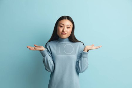 Photo for Confused Woman Shrugging Hands. Portrait of Uncertain Asian Girl Expressing Doubts and Bewilderment, Looking at Camera with Question So What, Who Cares. Indoor Studio Shot Blue Background - Royalty Free Image