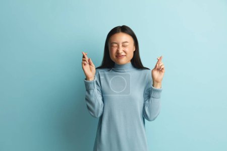 Photo for Asian Woman Clenched Fingers. Portrait of Hopeful Joyous Woman Raising Fingers Crossed While Making Wish, Confident to Win. Indoor Studio Shot Isolated on Blue Background - Royalty Free Image