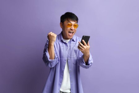 Photo for Winner Man Rejoicing Smartphone. Extremely Happy Asian Guy Raised Arm Gesturing Yes I Did It, Im Winner, Feeling Energetic and Lively. Isolated on Violet Background - Royalty Free Image