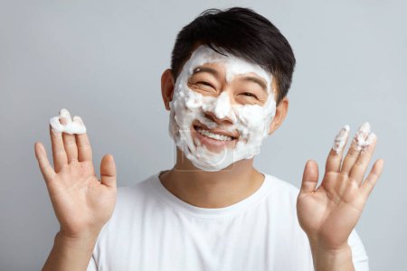 Foto de Tratamiento facial. Modelo asiático cara de limpieza con espuma blanca durante la relajación del spa. Cosmetología para el cuidado facial. Procedimiento de cuidado de la piel para el hombre Concepto - Imagen libre de derechos