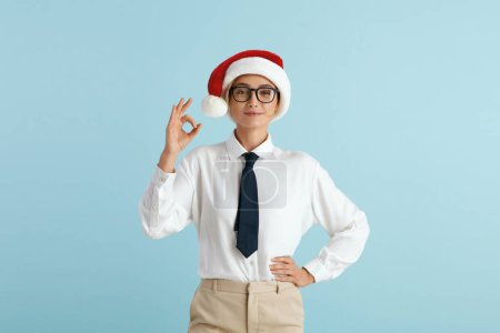 Photo for Smiling Businesswoman Showing Ok Gesture. Satisfied Caucasian Girl in Santa Hat Making Ok Symbol with Fingers, Approving, Satisfied. Indoor Studio Shot Isolated on Blue Background - Royalty Free Image