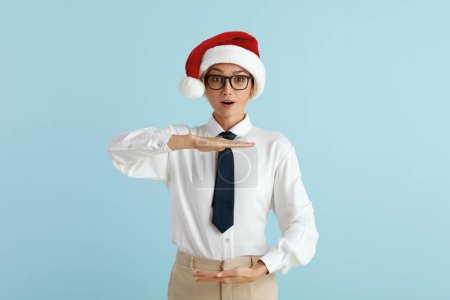 Photo for Surprised Businesswoman Showing Big Size. Satisfied Caucasian Lady in Santa Hat Showing Size With Both Hands, Looking at Camera With Surprised Expression. Indoor Shot Isolated on Blue Background - Royalty Free Image