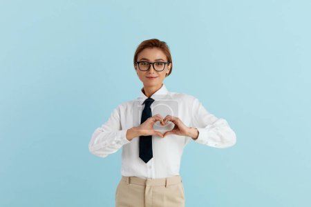 Photo for Businesswoman Showing Love Gesture. Portrait of Attractive Romantic Woman Standing Making Heart with Hands, Smiling Playfully. Indoor Studio Shot Isolated on Blue Background - Royalty Free Image