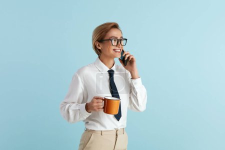 Photo for Busy Businesswoman Holding Coffee. Portrait of Friendly Young Woman in Glasses Talking Mobile, Holding Drink in Cup and Smiling. Indoor Studio Shot Isolated on Blue Background - Royalty Free Image