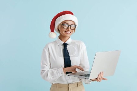 Téléchargez les photos : Joyeux Noël Businesswoman tenant ordinateur portable isolé. Femme blanche dans Santa Hat Ordinateur de travail. Lady Boss caméra souriante à fond bleu - en image libre de droit
