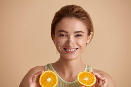 Foto de Laughing Girl Hold Slices of Orange. Mujer joven feliz mirando la cámara y la vitamina recomendada para la piel. Concepto de cuidado de la piel. Aislado sobre fondo beige - Imagen libre de derechos