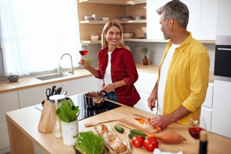 Téléchargez les photos : Couple cuisiner à la cuisine. Homme positif senior coupant des légumes biologiques frais pour la salade pendant que sa femme boit du vin. Une alimentation saine sur le concept de week-end romantique - en image libre de droit