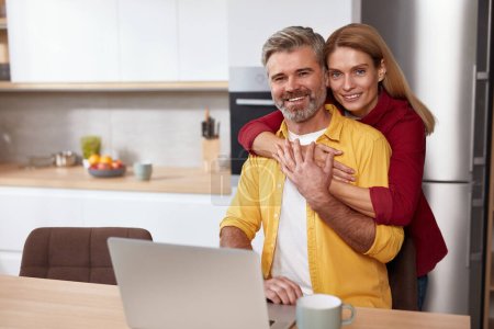 Happy Couple Embracing At Kitchen. Middle Aged Spouses Cuddling At Home Kitchen During The Morning. Wife Sitting In Front Of The Laptop 