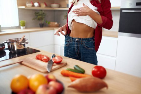 Téléchargez les photos : Diet Woman montrant combien de poids elle a perdu. Cropped View Of Diet Lady en jeans posant à la cuisine. Mode de vie sain concept - en image libre de droit