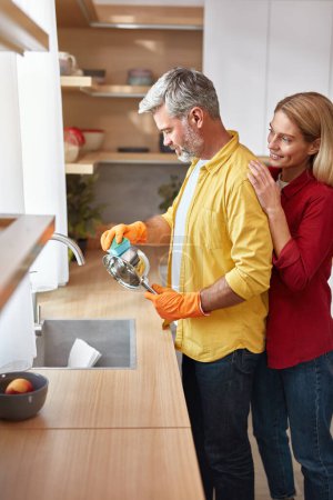Photo for Senior Couple Cleaning Dishes At Kitchen. Vertical Shot of Man Washing Dishes with Cleaning Liquid Under Tap Water. Cute Woman Embracing Husband in Modern Kitchen - Royalty Free Image