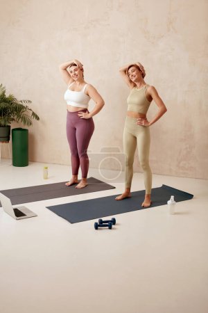 Photo for Positive Women Training Yoga. Girls In Sportswear Exercising At Home, Standing And Stretching Head On Fitness Mat, Full Length Shot. Healthy, Sporty Lifestyle, Yoga Practicing At Home - Royalty Free Image