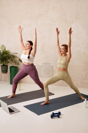 Photo for Yoga Women Training Together. Girls In Sportswear Exercising At Home, Standing In Warrior Pose On Fitness Mat, Full Length Shot. Healthy, Sporty Lifestyle, Yoga Practicing At Home - Royalty Free Image