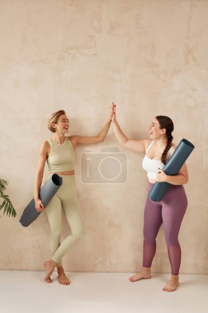 Foto de Mujer sonriente dando cinco después del yoga. Mujeres amigas riendo y sosteniendo colchonetas de yoga después de la sesión de yoga juntas en casa. Chicas atractivas en ropa deportiva pasar tiempo libre de ocio - Imagen libre de derechos