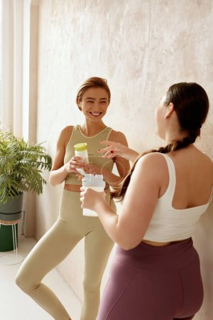 Téléchargez les photos : Femme souriante qui parle après le yoga. Des amies riant et tenant des boissons de désintoxication après une séance de yoga ensemble à la maison. Filles attrayantes en vêtements de sport passer du temps libre de loisirs - en image libre de droit