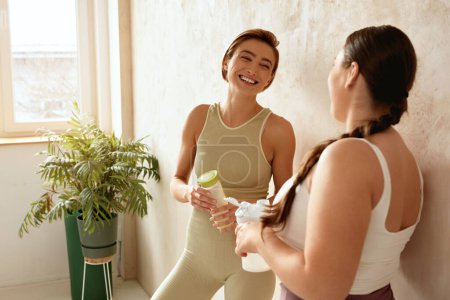 Téléchargez les photos : Femme souriante qui parle après le yoga. Des amies riant et tenant des boissons de désintoxication après une séance de yoga ensemble à la maison. Filles attrayantes en vêtements de sport passer du temps libre de loisirs - en image libre de droit