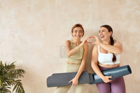 Téléchargez les photos : Femmes riantes posant après le yoga. Amis féminins souriant et montrant geste de coeur après la séance de yoga ensemble à la maison. Filles attrayantes en vêtements de sport passer du temps libre de loisirs - en image libre de droit