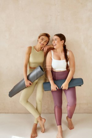 Foto de Mujeres riéndose abrazando después del yoga. Mujeres amigas riendo y sosteniendo colchonetas de yoga después de la sesión de yoga juntas en casa. Chicas atractivas en ropa deportiva pasar tiempo libre de ocio - Imagen libre de derechos