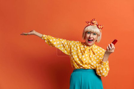 Foto de Feliz abuela sosteniendo móvil. Imagen de una mujer mayor de pie sobre un fondo naranja aislado, mirando a la cámara, sonriendo y sintiéndose feliz - Imagen libre de derechos