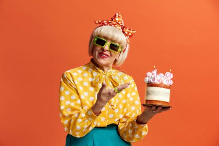Téléchargez les photos : Joyeux anniversaire de grand-mère. Amusé à la mode Senior Lady célèbre anniversaire tient gâteau avec des bougies brûlantes isolées sur fond orange. Positive mamie célèbre Bday - en image libre de droit