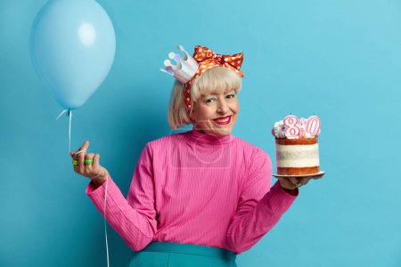 Happy Grandmother Celebrating Birthday. Positive Senior Lady Posing With Cake And Inflated Balloon Isolated Over Blue Background. Positive Granny Smiling Broadly Having Festive Mood During Party