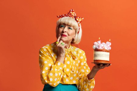 Téléchargez les photos : Méditer grand-mère fête son anniversaire. Amusé à la mode Senior Lady célèbre anniversaire tient gâteau avec des bougies brûlantes isolées sur fond orange. Positive mamie célèbre Bday - en image libre de droit
