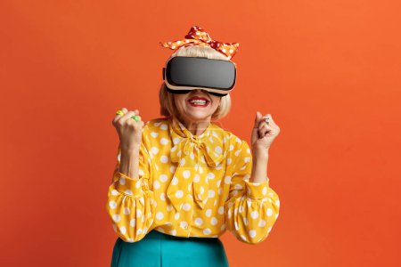 Foto de Mujer emocionada probando VR. Retrato de la abuela mayor sorprendida Descubriendo nuevas tecnologías con auriculares de realidad virtual, visión 3D futurista. Tiro de estudio interior aislado sobre fondo naranja - Imagen libre de derechos