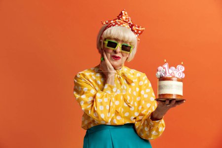Téléchargez les photos : Joyeux anniversaire de grand-mère. Amusé à la mode Senior Lady célèbre anniversaire tient gâteau avec des bougies brûlantes isolées sur fond orange. Positive mamie célèbre Bday - en image libre de droit