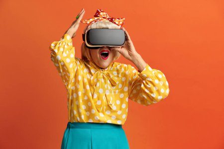 Foto de Mujer emocionada probando VR. Retrato de la abuela mayor sorprendida Descubriendo nuevas tecnologías con auriculares de realidad virtual, visión 3D futurista. Tiro de estudio interior aislado sobre fondo naranja - Imagen libre de derechos