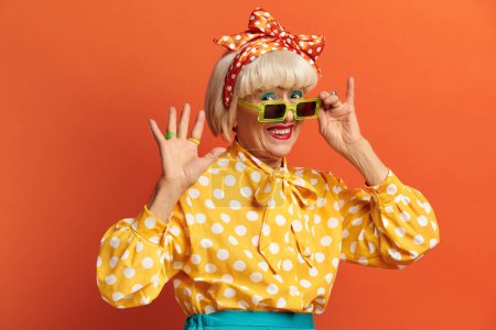Photo for Coquettish Grandmother Gesturing Hello. Beautiful Mature Woman Lift Her Arm Looking Straight In Camera With Happy Smile Isolated On Orange Background - Royalty Free Image