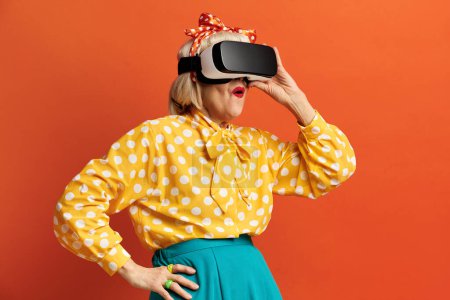 Téléchargez les photos : Femme excitée essayant VR. Portrait d'une grand-mère aînée étonnante découvrant les nouvelles technologies portant un casque de réalité virtuelle, vision 3D futuriste. Studio intérieur isolé sur fond orange - en image libre de droit