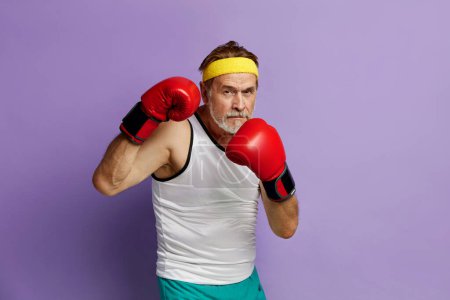 Foto de Hombre fuerte boxeando en guantes. Retrato de un hombre mayor entrenando y demostrando poder en sus manos, sintiendo energía en su época. Estudio interior filmado aislado sobre fondo violeta - Imagen libre de derechos