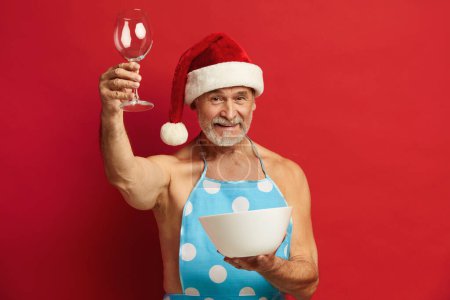 Téléchargez les photos : Homme aîné tenant des biceps en verre à vin. Homme barbu dans Santa Chapeau et tablier sur torse nu posant avec plaque et verre isolé sur fond rouge Studio - en image libre de droit