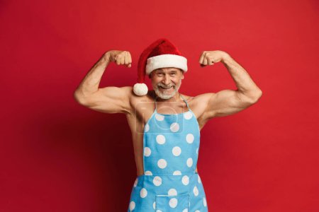 Photo for Senior Man Showing Biceps. Strong Bearded Man In Santa Hat And Apron On Naked Torso Posing Isolated On Red Background Studio. Showing Biceps, Muscles - Royalty Free Image