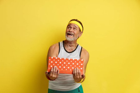 Téléchargez les photos : Senior Man Holding Present. Portrait de beau barbu grand-père posant avec boîte-cadeau avec expression faciale curieuse, cadeau d'anniversaire. Studio intérieur Shot isolé sur fond jaune - en image libre de droit