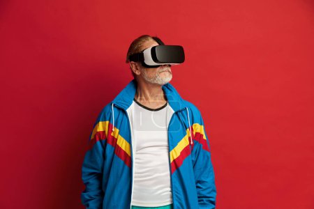Téléchargez les photos : Focused Man Essayant VR. Portrait de l'homme âgé Découvrir les nouvelles technologies Porter un casque de réalité virtuelle, Vision 3D futuriste. Studio intérieur isolé sur fond rouge - en image libre de droit