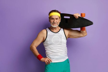 Téléchargez les photos : Un homme souriant tenant du skateboard. Blanc senior homme tenant Longboard sur épaule et souriant tout en posant isolé sur fond violet - en image libre de droit