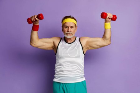 Photo for Strong Man Training With Dumbbells. Glad Grandfather Raising Arms With Dumbbells, Satisfied After Having Training In Gym Isolated Over Purple Wall Empty Space - Royalty Free Image