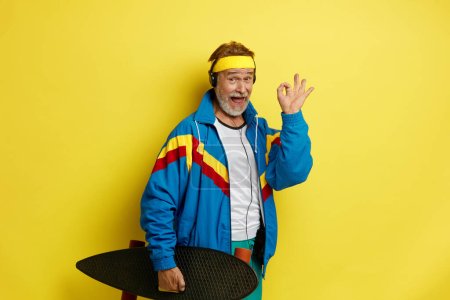 Photo for Smiling Man Holding Skateboard. Headphones Senior Man Showing Ok Gesture With Longboard on Shoulder and Listening Music While Posing Isolated on Yellow Background - Royalty Free Image
