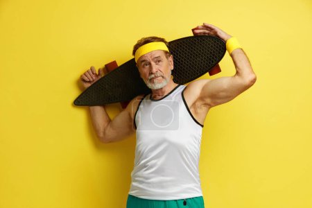 Téléchargez les photos : Un homme souriant tenant du skateboard. Blanc senior homme tenant Longboard sur épaule et souriant tout en posant isolé sur fond jaune - en image libre de droit