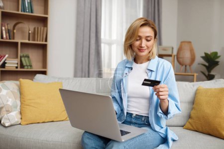 Foto de Positive Woman Holding Credit Card. Smiling Woman Paying Online, Using Laptop, Holding Plastic Credit Card, Sitting On Couch At Home. Young Female Shopping, Making Secure Internet Payment - Imagen libre de derechos