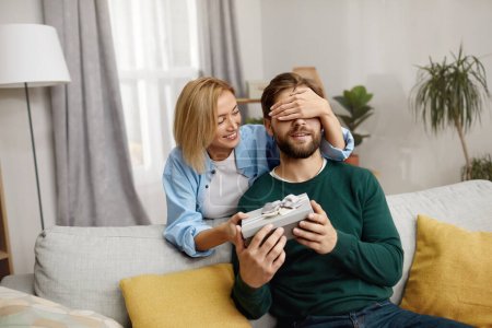 Photo for Happy Woman Presenting Gift To Husband. Excited Young Woman Closed Eyes Presenting Good Unexpected Present To Husband At Home. Loving Girlfriend Making Romantic Surprise To Boyfriend - Royalty Free Image