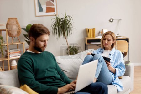Photo for Couple Using Gadgets At Home. Married Couple Of Freelancers Work From Home. Guy And Girl In Casual Clothes With Gadgets Work In The Living Room At The Sofa - Royalty Free Image