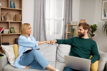 Téléchargez les photos : Couple Using Gadgets At Home. Married Couple Of Freelancers Bumping Hands While Work From Home. Guy And Girl In Casual Clothes With Gadgets Work In The Living Room At The Sofa - en image libre de droit
